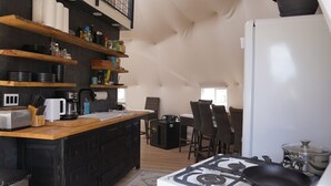 Een compacte keuken met een gasfornuis, een koffiezetapparaat en een broodrooster. Er staan rieten stoelen bij een kleine eettafel.