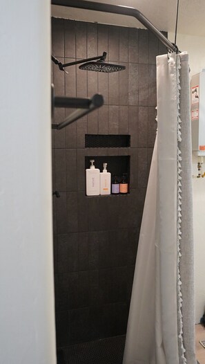Un espace douche avec un mur recouvert de carreaux foncés, une pomme de douche et une étagère contenant des articles de toilette.