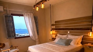 A bedroom with a bed, bedside lamps, a bench, a chair, a window with curtains, and a view of the sea. - Zephyrus Sunset House  (Naxos)
