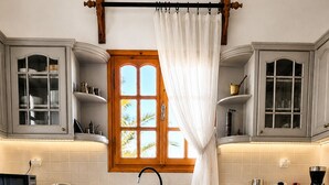 A kitchen with a central island, a window with a curtain, and built-in cabinets. - Zephyrus Sunset House  (Naxos)