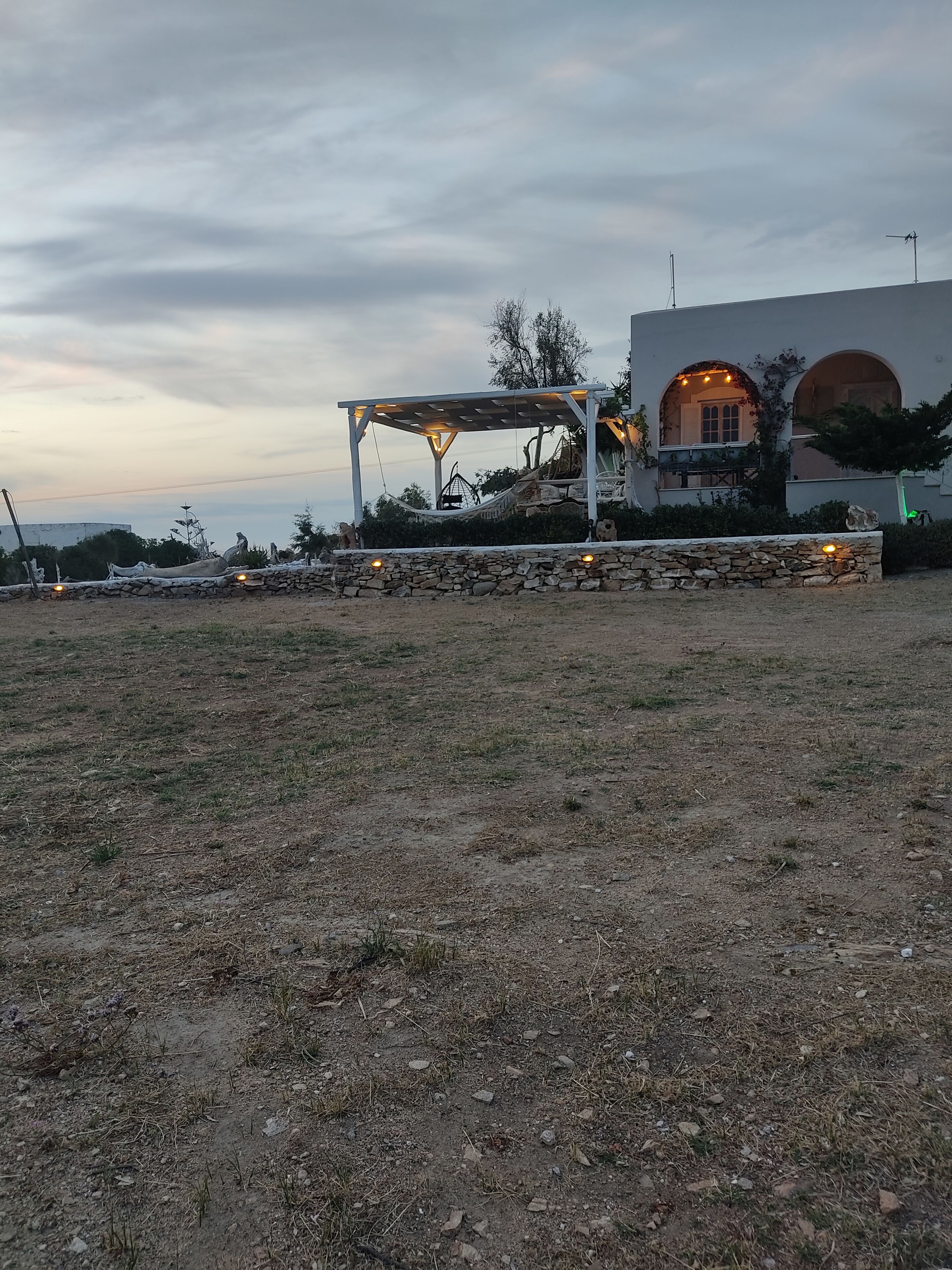 A stone wall with a pergola and string lights, a stone pathway, and a building with arches and a balcony.