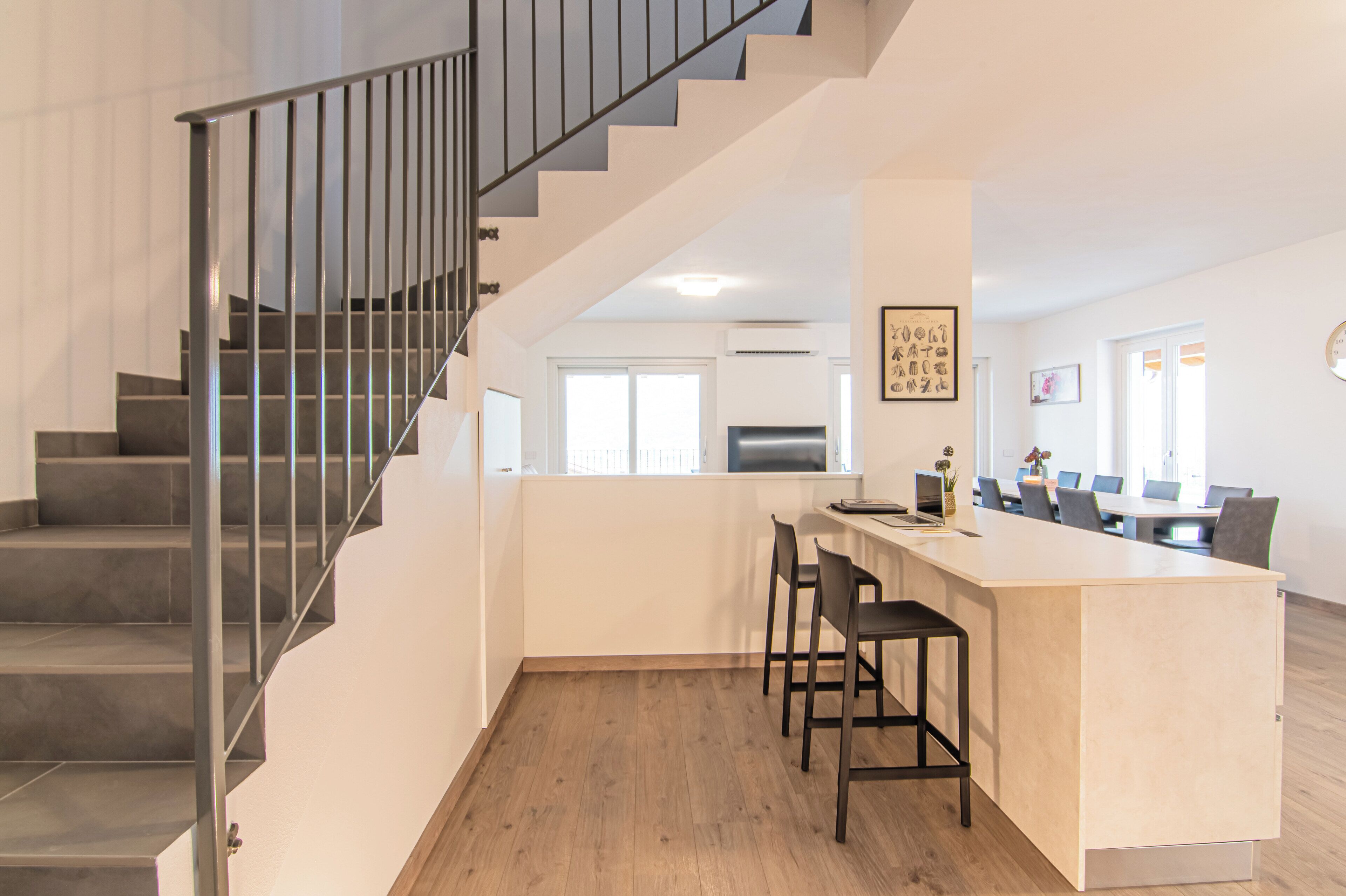 A modern kitchen with a central island, a dining area with chairs, and a staircase with metal railings.