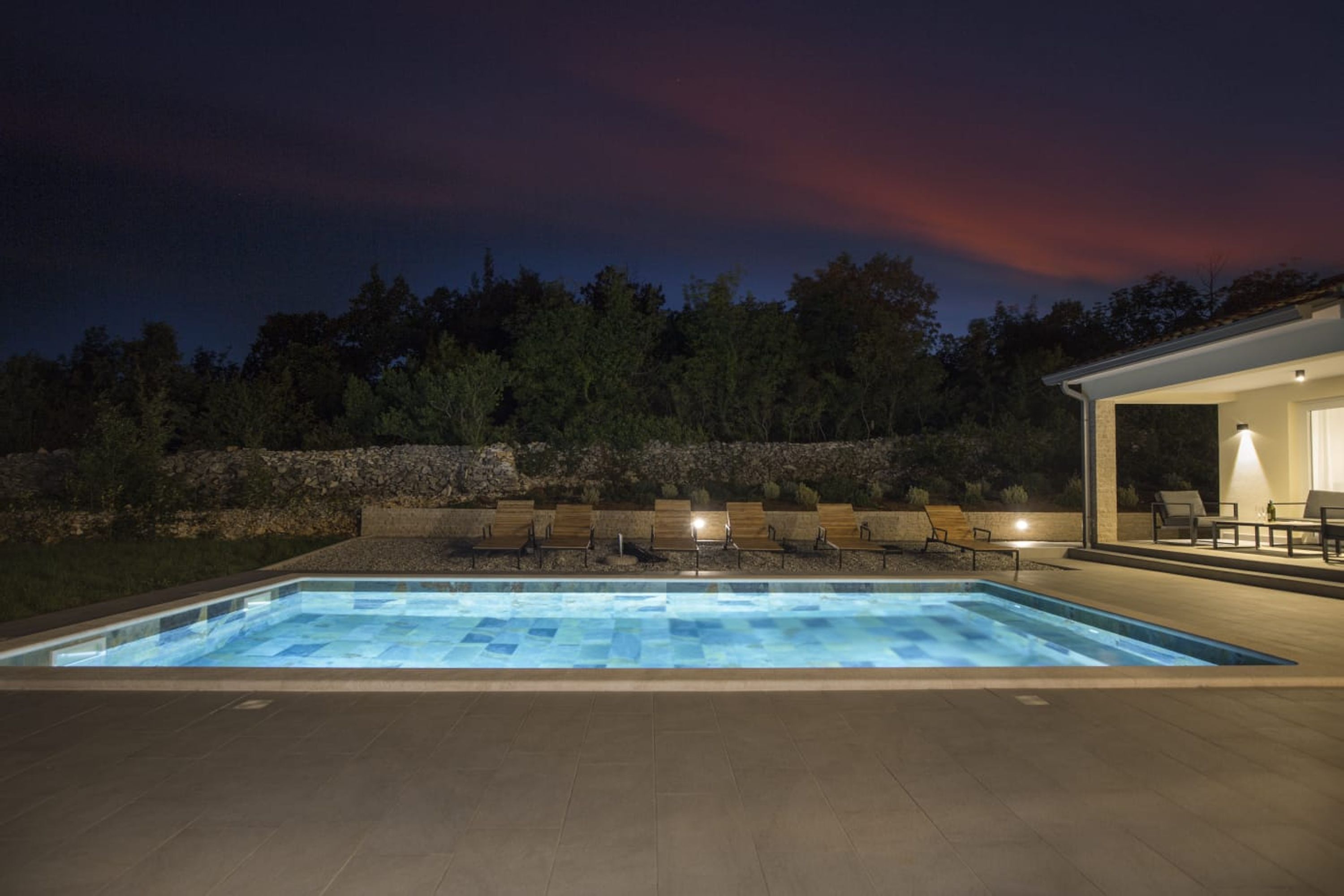 A rectangular swimming pool with a clear blue water feature, surrounded by a stone wall and a seating area with chairs.