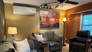 Una sala de estar con un sofá, un sillón y una mesita. Hay un ventilador de techo, un equipo de aire acondicionado y una lámpara de pie. En la pared, un cuadro de un paisaje urbano.