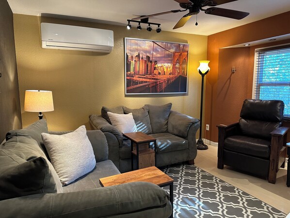 A living room with a sofa, armchair, and a small table. There is a ceiling fan, an air conditioning unit, and a floor lamp. A cityscape painting is on the wall. - Cozy 1-bedroom suite on a small goat farm with full kitchen and living room (Port Orchard)