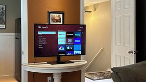Un televisor TCL Roku sobre una mesa blanca tipo pedestal en una sala de estar, con una pared marrón y una puerta al fondo.