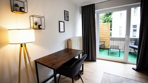 Una habitación moderna con una mesa de madera, una silla negra y una lámpara de pie. Se ve una ventana con vistas a un jardín y un edificio.
