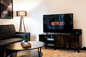 A living room with a sofa, a coffee table with a plant, a TV displaying 'NETFLIX', a lamp, and a wooden cabinet with a game console.