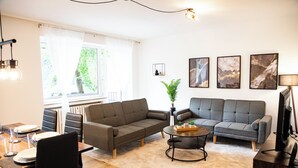 Una sala moderna con dos sofás grises, una mesa de centro y una zona de comedor con mesa y sillas. Hay obras de arte enmarcadas en la pared y una planta en maceta en la esquina.