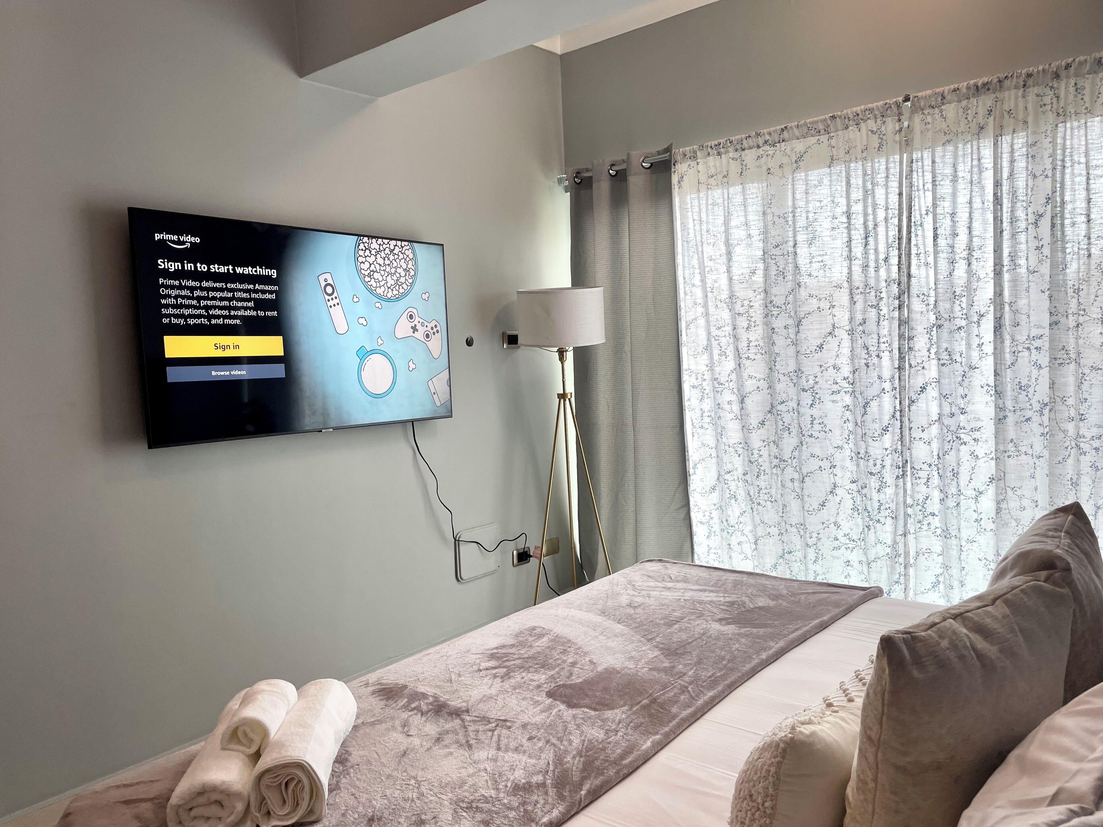 Ett hotellrum med en säng, en platt-TV som är monterad på väggen, en lampa och ett fönster med blommiga gardiner.