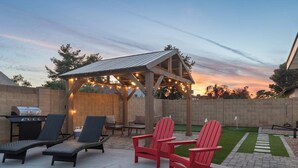 Un jardin avec un barbecue, des chaises longues et un kiosque illuminé de guirlandes lumineuses.