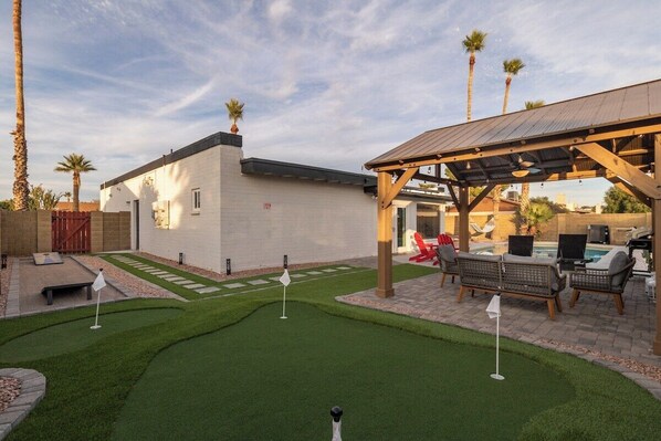 A backyard with a putting green, patio furniture, and a covered seating area.
