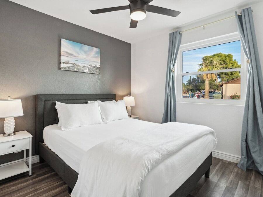 Un dormitorio ordenado con cama, mesitas de noche, ventilador de techo y ventana con cortinas.