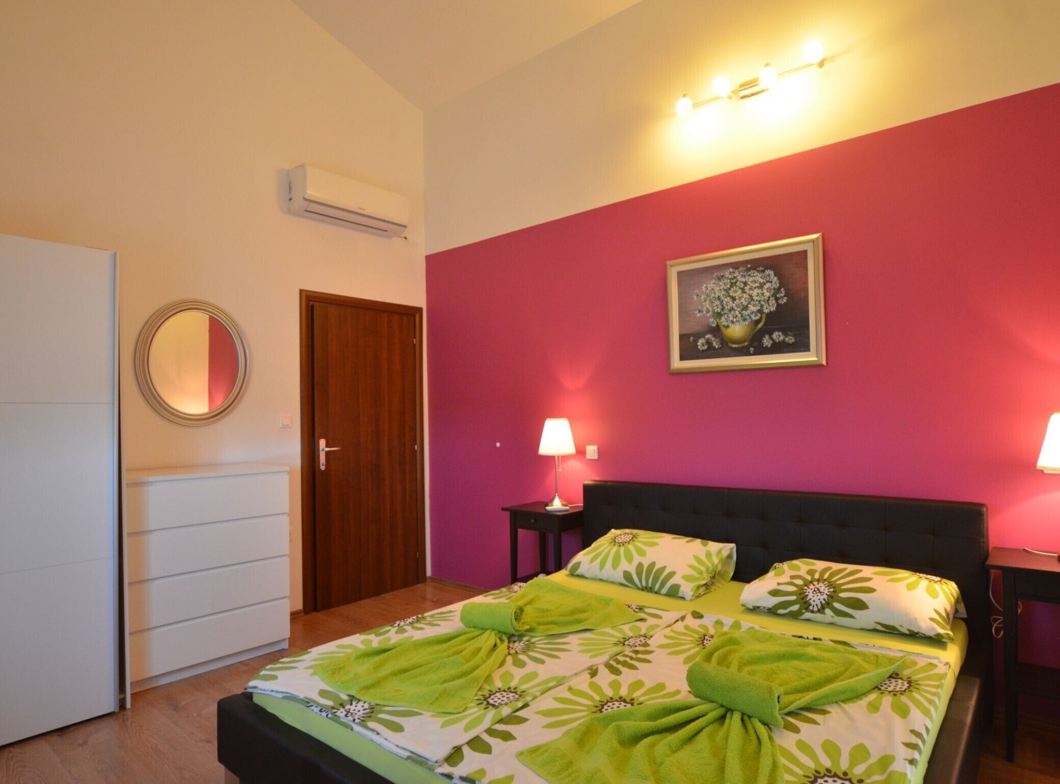A bedroom with a bed, two bedside lamps, a door, an air conditioner, and a framed picture on the wall.