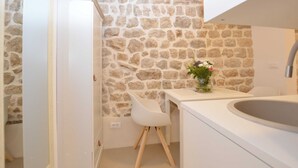 Une salle de bain avec un mur en pierre, un lavabo blanc, une chaise blanche et un vase rempli de fleurs.