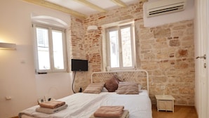 A bedroom with a stone wall, a bed with striped bedding, a flat-screen TV, and a small bedside table. - Modern and Cozy Rovinj Old Town Apartments - Classic Studio Apartment (Rovinj)