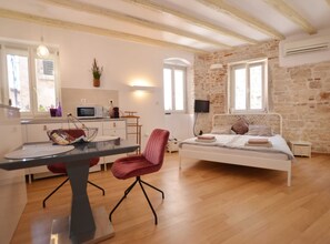 Une chambre avec un lit, un bureau et une chaise. Il y a un coin cuisine équipé d’un four à micro-ondes et d’un évier. Les murs de la pièce sont en pierre et le plafond est orné de poutres en bois.