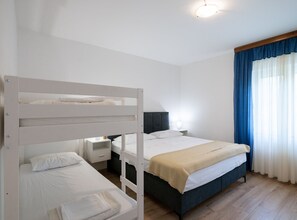 Une chambre avec un lit superposé, un lit simple, une table de chevet avec une lampe et une fenêtre avec des rideaux bleus.