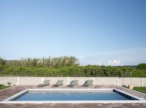 Une piscine rectangulaire bordée de quatre chaises longues disposées en ligne le long d’un de ses côtés.