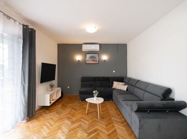 Un salon moderne avec un canapé gris, une table d’appoint blanche et un parquet en bois à motifs chevrons.