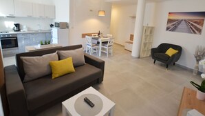 A modern living room with a brown sofa, a white coffee table, and a remote control on it. There is a dining area with a white table and chairs, and a kitchen with white cabinets and appliances. - Seaside Escape Rovinj Old Town Apartment - Three Bedroom Apartment with Sea View (Rovinj)
