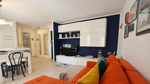 Ein modernes Wohnzimmer mit einem orangefarbenen Sofa, einem Essbereich mit weißem Tisch und Stühlen sowie einer Küchenzeile mit weißen Schränken und einem Fernseher.