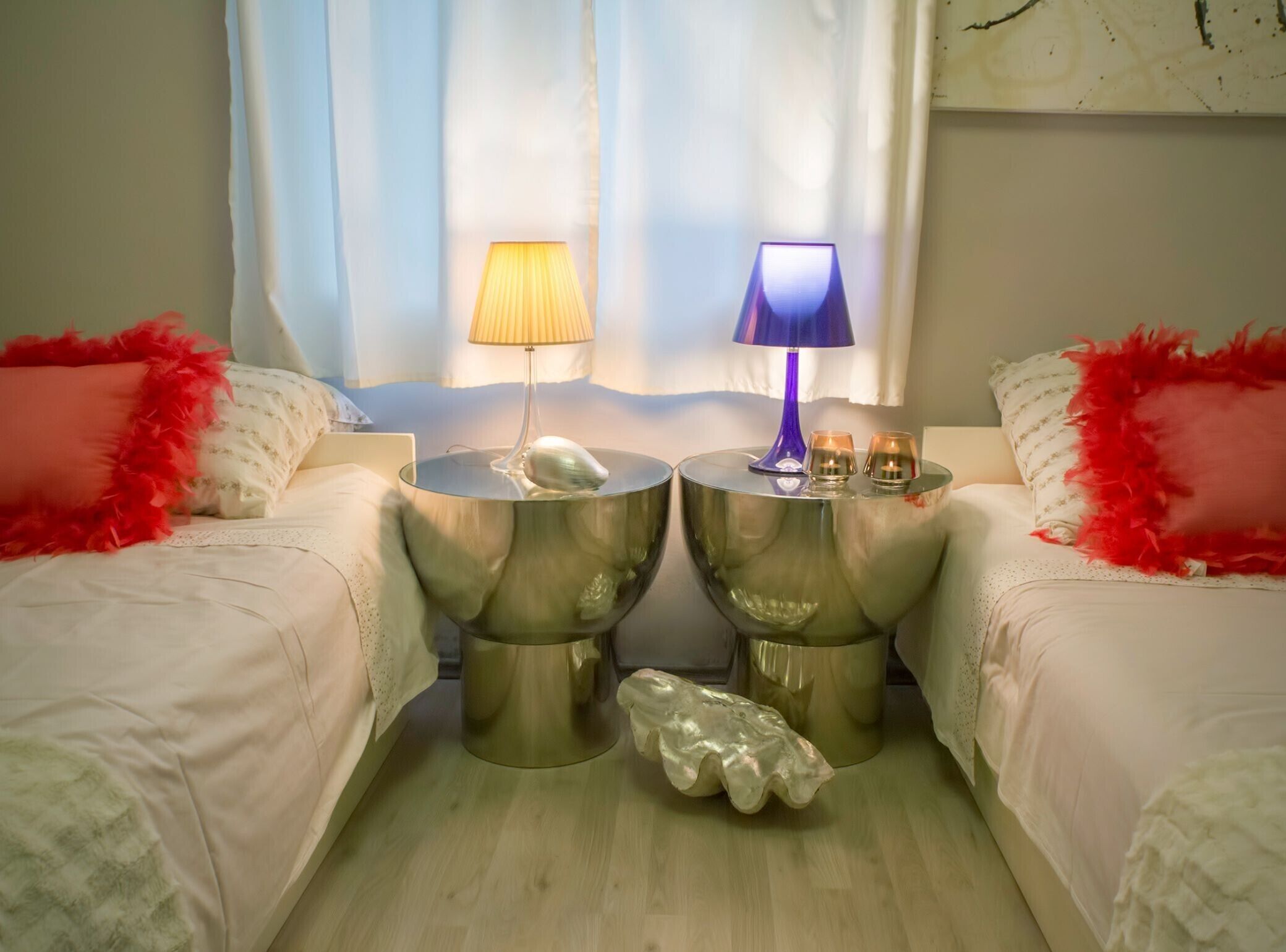 A hotel room with two beds, each with a red feathered pillow, a round side table with a lamp, and a decorative shell.