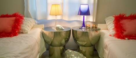 A hotel room with two beds, each with a red feathered pillow, a round side table with a lamp, and a decorative shell.