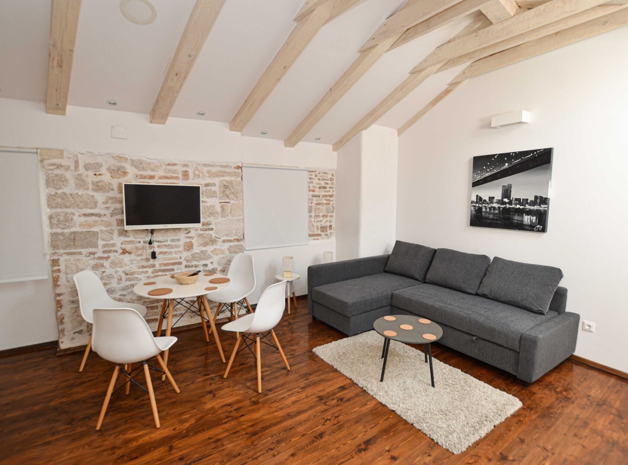 Una sala de estar moderna con una pared de piedra, un sofá gris, sillas blancas, una mesa redonda y una pintura de un paisaje urbano.