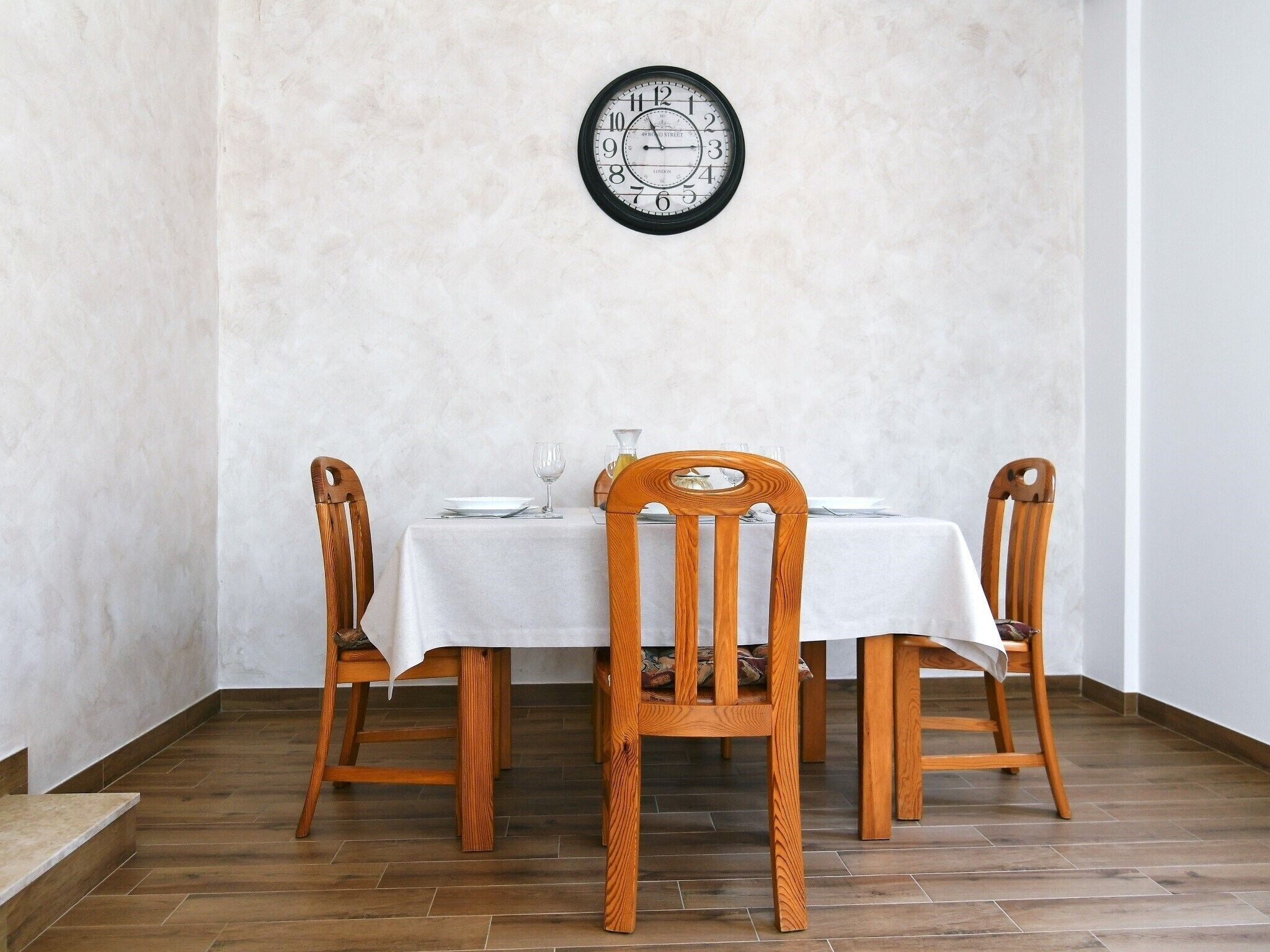 Un comedor con una mesa dispuesta para dos personas, sillas de madera, un reloj de pared y un mantel blanco.
