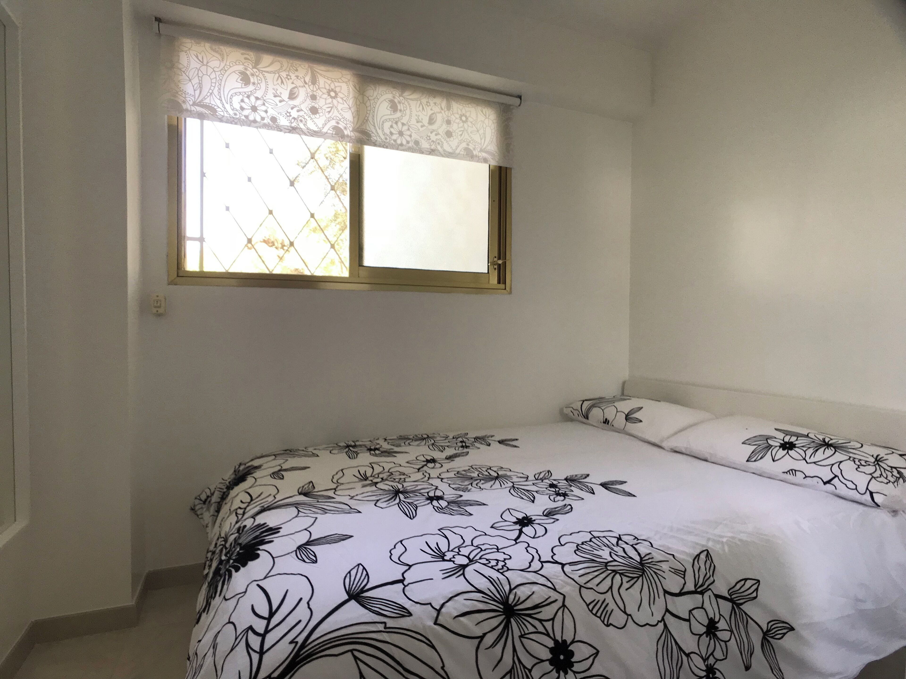 A bedroom with a bed, a window with a patterned curtain, and a plain wall.