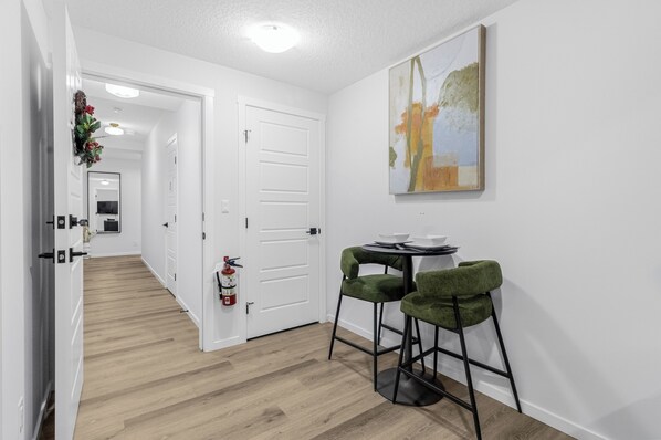 Un couloir avec un plancher en bois, des murs blancs, une porte et un extincteur. On y trouve une petite table avec deux chaises vertes et un tableau accroché au mur.
