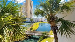 A view of a hotel courtyard with a swimming pool, palm trees, and a tall building in the background. - Enjoy a spacious, low-rise Sugar Beach condo with a private beach entrance, pool, sunset views, and glimpses of the Gulf. The condo sleeps up to 10 and is close to dining and local attractions in Panama City Beach. (Panama City Beach)