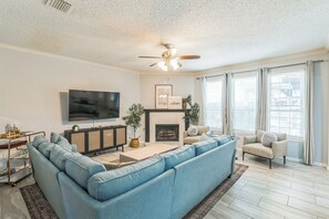 A modern living room with a sectional sofa, a flat-screen TV, a fireplace, and large windows with curtains. - Rockwall Retreat | Pool, Games & King Beds (Rockwall)