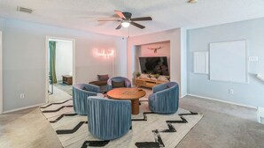 Un salon moderne doté d’un ventilateur de plafond, d’un grand téléviseur à écran plat et d’un coin salon confortable avec des fauteuils bleus et une table basse en bois.