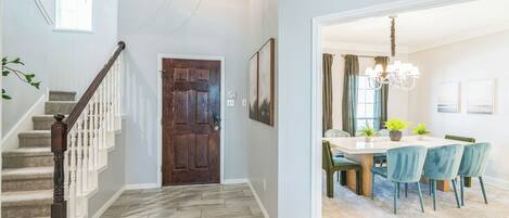 A hallway with a wooden door, a staircase with white balusters, and a dining area with a chandelier.