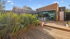 Een modern huis met een houten terras, een glazen deur naar een buiten eethoek, en een tuin met tropische planten.
