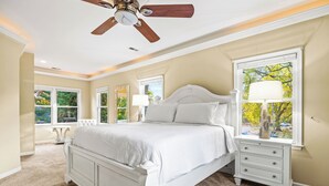 Une chambre à coucher avec un grand lit, une table de chevet, un ventilateur de plafond et une fenêtre donnant sur des arbres.