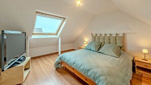 Quarto com teto inclinado, claraboia, cama com colcha clara, mesa de cabeceira com abajur e TV sobre suporte de madeira.