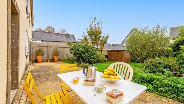 Um pátio com uma mesa posta para o café da manhã, incluindo pão, bananas, uma cafeteira e uma xícara.