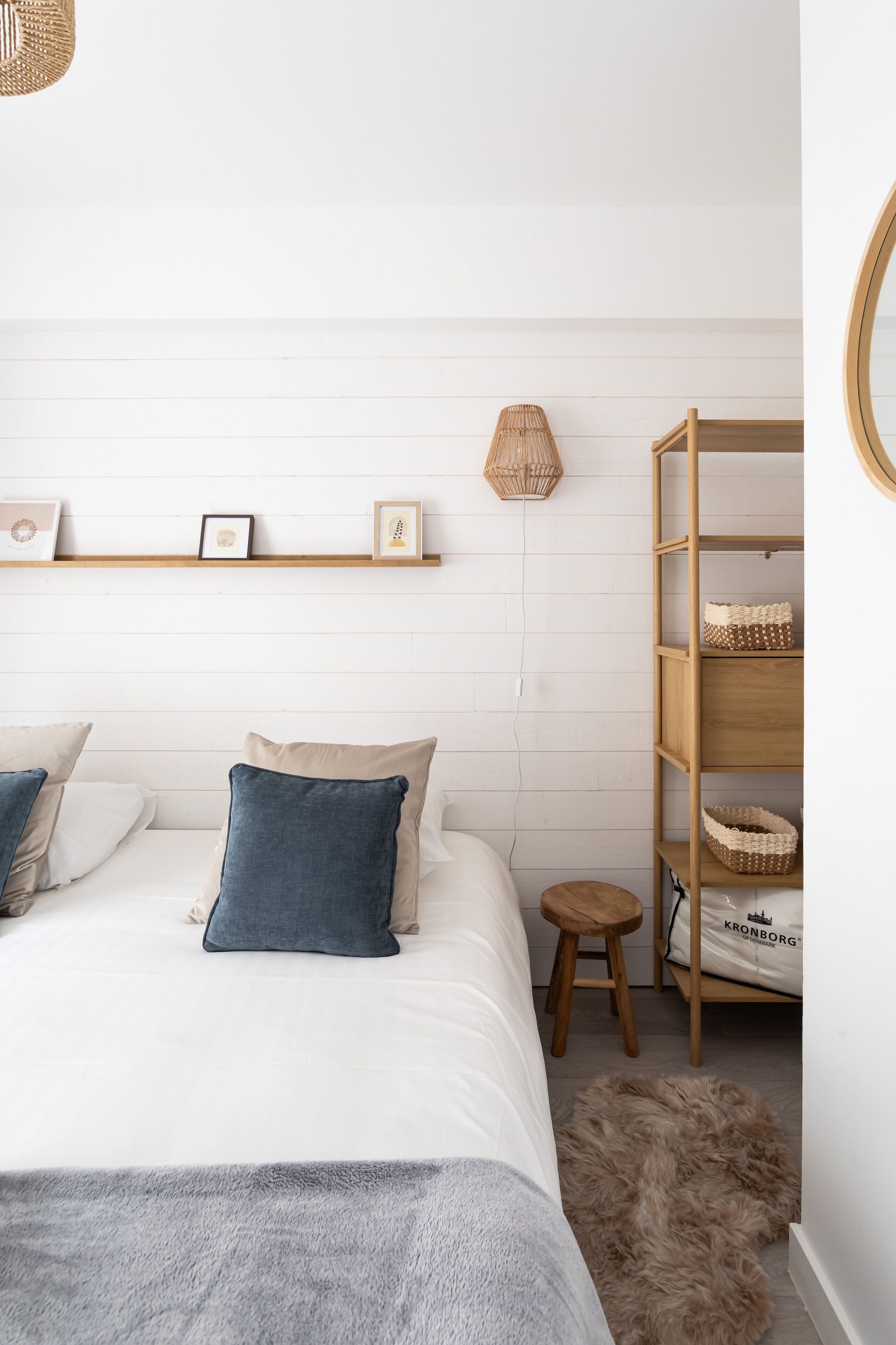 Une chambre à coucher bien rangée, avec un lit, des oreillers, un petit tabouret, une étagère avec des photos encadrées et un panier en osier posé sur une étagère en bois.
