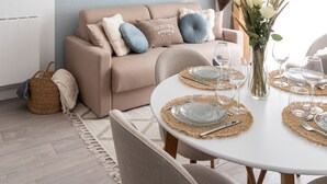 Ein modernes Wohnzimmer mit einem beigen Sofa, einem runden Esstisch für vier Personen und einem großen Fenster mit Vorhängen.