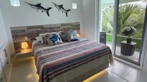 A bedroom with a bed, a ceiling fan, two wall-mounted fish decorations, a nightstand with a lamp, and a view through a sliding glass door to a balcony with a potted plant. - COCO SAI, where you will see the majesty of the sea and the sky as one.  (San Andrés)