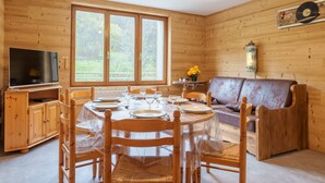A wooden cabin interior with a dining table set for four, a sofa, a television, and a large window. - Les Jonquilles - 3 chambres et garage privé (Cornimont)