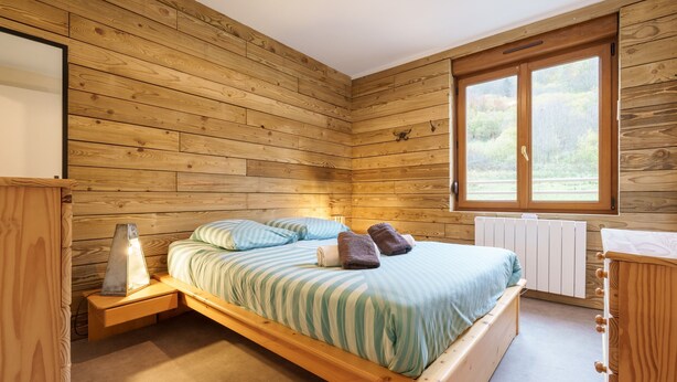 A wooden-paneled bedroom with a bed, bedside table, and a window with a view of greenery. - Les Jonquilles - 3 chambres et garage privé (Cornimont)