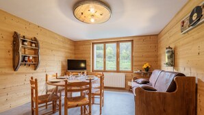 A wooden interior with a dining table, chairs, a sofa, a TV, and a window with a view of greenery. - Les Jonquilles - 3 chambres et garage privé (Cornimont)