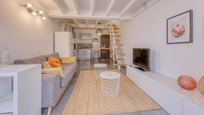 A modern living room with a grey sofa, white coffee table, and a TV. There is a kitchen in the background with a white fridge and a dining area with stools.