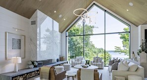 A spacious living room with a large window offering a view of trees and water. There is a fireplace, comfortable seating, and a modern chandelier.