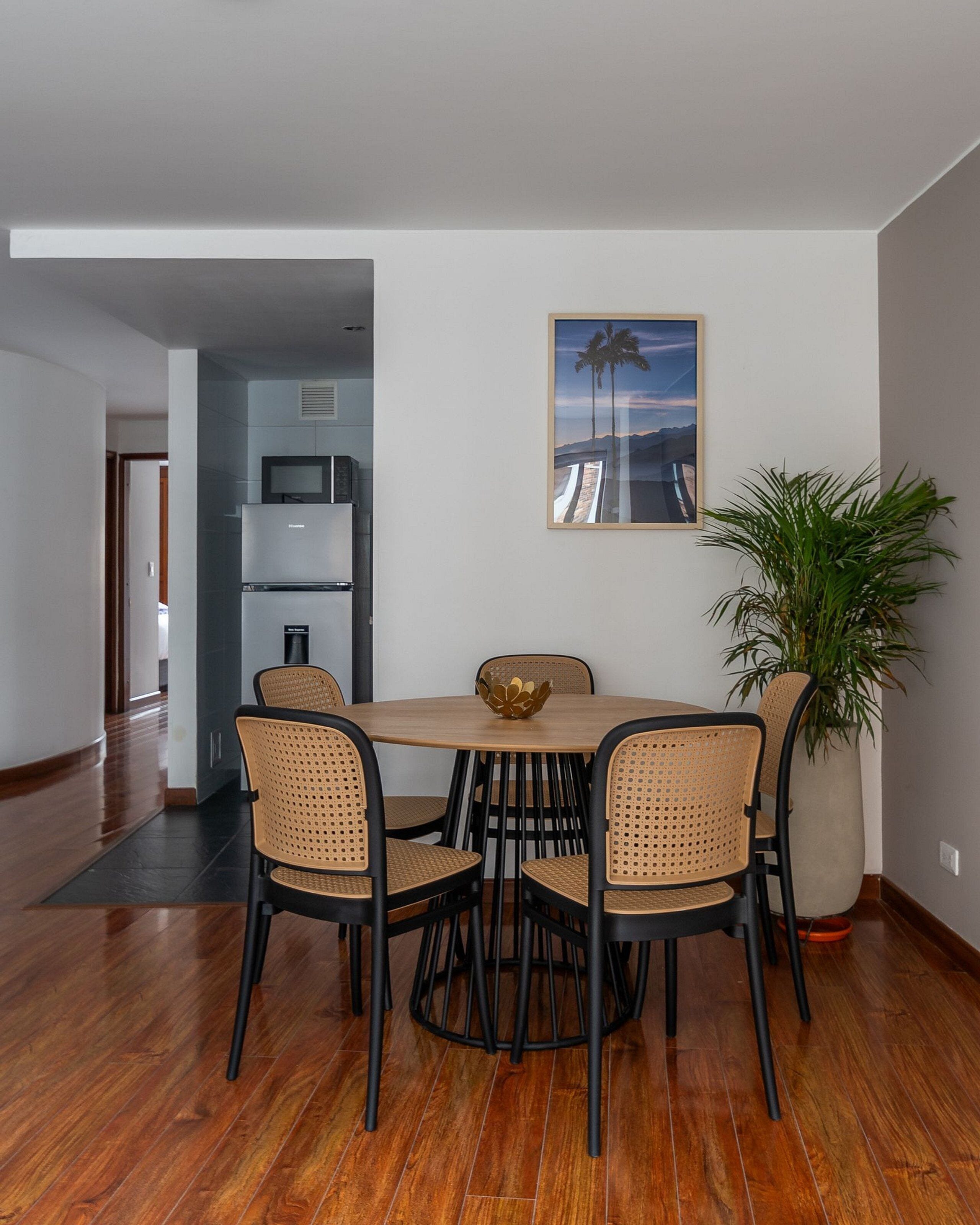 A dining area with a wooden table and four chairs, a large potted plant, and a framed picture on the wall.