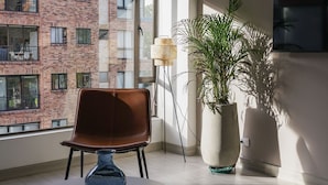 A modern living room with a large window, a brown chair, a round table, and two potted plants. - 10.73 Flats x Ahead (Bogotá)